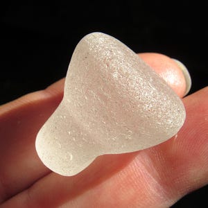May include: A piece of translucent, frosted white sea glass with a smooth, rounded, and slightly tapered shape. The surface of the glass has a weathered, matte texture with small indentations, giving it a soft, diffused appearance.