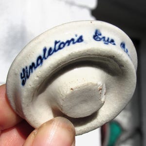 May include: A small, off-white ceramic bottle stopper with a rounded top and a short, cylindrical base. The text "Singleton's Eye" is written in blue script around the edge of the stopper. The stopper has a slightly aged appearance.