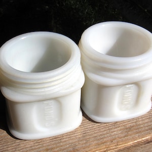 May include: Two white milk glass jars with open tops and threaded rims. Each jar features a rectangular recessed panel on the front with the embossed text POND'S. The containers have a rounded square base and a smooth, opaque finish throughout.
