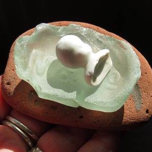 May include: A miniature white ceramic jug sits on a piece of pale green sea glass, which is set on a reddish-brown stone. The stone has a smooth, rounded shape. The background is dark.