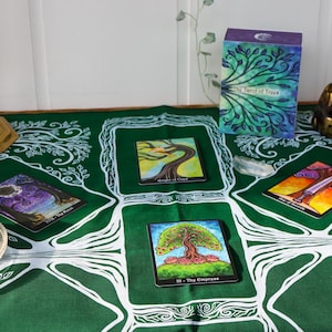 May include: A green tarot cloth with white tree designs, featuring tarot cards with colorful illustrations. A deck of cards, a box, and a golden Buddha statue are also present. A brass candle holder and a bowl with herbs are on the table.