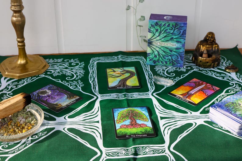 May include: A green tarot cloth with white tree designs, featuring tarot cards with colorful illustrations. A deck of cards, a box, and a golden Buddha statue are also present. A brass candle holder and a bowl with herbs are on the table.
