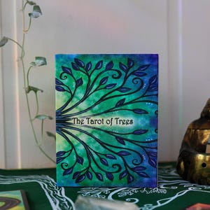 May include: A tarot card deck titled "The Tarot of Trees." The deck features a watercolor design of tree branches with blue leaves against a green and blue background. The text is in a white, stylized font.