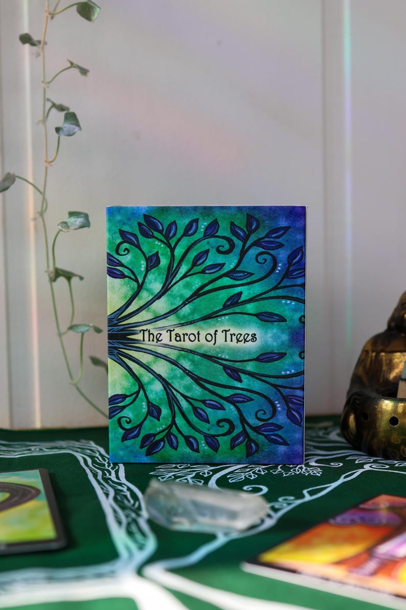 May include: A tarot card deck titled "The Tarot of Trees." The deck features a watercolor design of tree branches with blue leaves against a green and blue background. The text is in a white, stylized font.