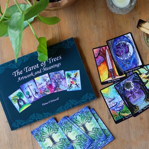 May include: A collection of tarot cards and a book titled "The Tarot of Trees: Artwork and Meanings" are arranged on a wooden surface. The book is dark teal with colorful illustrations. Several tarot cards are displayed, featuring vibrant artwork and imagery.