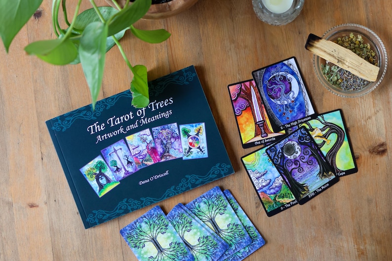 May include: A collection of tarot cards and a book titled "The Tarot of Trees: Artwork and Meanings" are arranged on a wooden surface. The book is dark teal with colorful illustrations. Several tarot cards are displayed, featuring vibrant artwork and imagery.