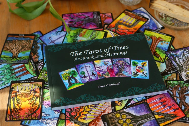 May include: A collection of colorful tarot cards and a book titled "The Tarot of Trees: Artwork and Meanings" by Dana O'Driscoll. The cards feature vibrant illustrations of trees and nature scenes, with a dark green book cover.