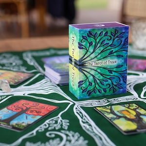 May include: A tarot card deck titled "The Tarot of Trees" with a watercolor design of a tree on the box. The box is on a green cloth with tree-themed illustrations and several tarot cards. A clear crystal is also on the cloth.