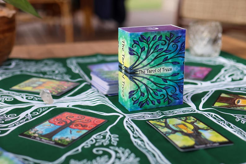 May include: A tarot card deck titled "The Tarot of Trees" with a watercolor design of a tree on the box. The box is on a green cloth with tree-themed illustrations and several tarot cards. A clear crystal is also on the cloth.