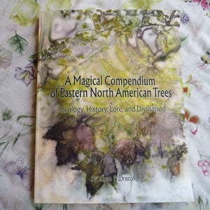 May include: A book titled "A Magical Compendium of Eastern North American Trees: Ecology, History, Lore, and Divination" by Dena Driscoll. The cover features a watercolor-style illustration of leaves in shades of green, brown, and purple.