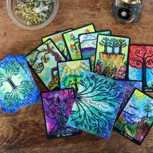 May include: A collection of tarot cards featuring colorful tree illustrations. The cards are arranged on a wooden surface, with a title card reading "The Tarot of Trees." The artwork includes vibrant blues, greens, reds, and purples. A small glass bowl and candle are also visible.