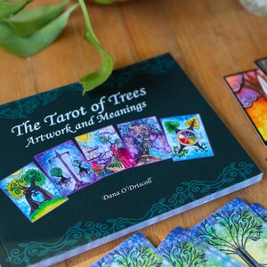 May include: A dark green book titled "The Tarot of Trees Artwork and Meanings" with colorful tarot cards. The book cover features several tarot card images. The cards are scattered around the book on a wooden surface.