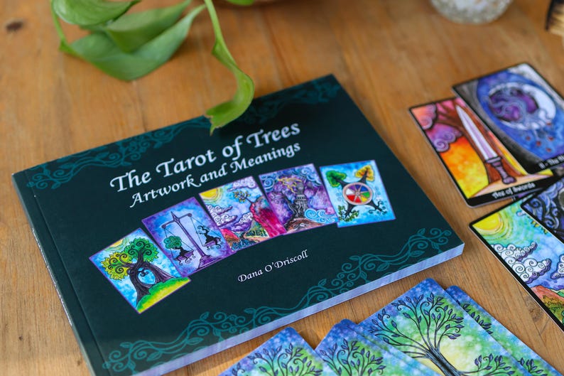 May include: A dark green book titled "The Tarot of Trees Artwork and Meanings" with colorful tarot cards. The book cover features several tarot card images. The cards are scattered around the book on a wooden surface.