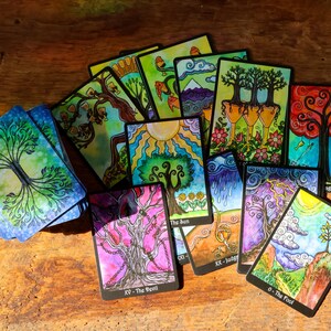 May include: A collection of colorful tarot cards featuring vibrant illustrations of trees, suns, and landscapes. The cards are arranged on a wooden surface, with titles such as "The Sun" and "The Devil" visible. The artwork is detailed and whimsical.