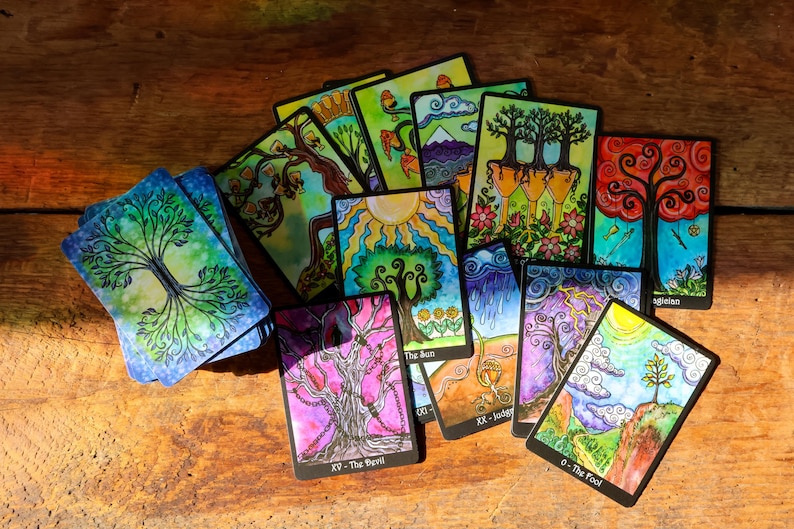 May include: A collection of colorful tarot cards featuring vibrant illustrations of trees, suns, and landscapes. The cards are arranged on a wooden surface, with titles such as "The Sun" and "The Devil" visible. The artwork is detailed and whimsical.