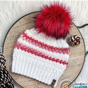 Può includere: Berretto in crochet bianco con strisce rosse e bianche e un pompon rosso soffice. Il berretto ha un piccolo cartellino marrone con un fiocco di neve.