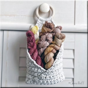 以下が含まれることがあります： 木製リングの上部に付いた、クロシェの吊り下げバスケット。黄色、ピンク、茶色、赤の色合いのカラフルな糸玉が入っています。