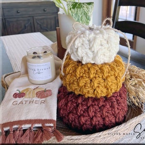 Pode incluir: Uma pilha de três abóboras de crochê em branco, amarelo mostarda e bordô. As abóboras estão em uma cesta de vime com uma toalha branca que diz "Gather" em bordado marrom.