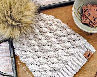 Puede incluir: Un gorro de punto blanco con un pompón de piel sintética marrón. El gorro está en una bandeja de madera con un cuenco marrón de pequeñas etiquetas de cuero que dicen "Shk". También hay un gancho de ganchillo en la bandeja.
