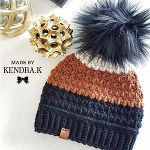 May include: A black, brown and cream striped crocheted beanie with a black faux fur pom pom. The beanie has a brown leather tag with the text "MADE BY KENDRA.K" on it.