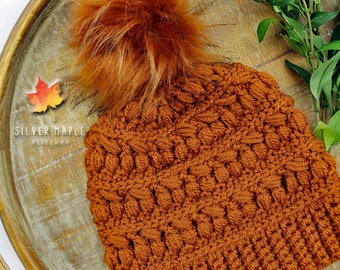 Op de afbeelding: Een gebreide beanie muts in een warme bruine kleur met een pluche bruine pompon bovenop. De muts ligt op een houten dienblad met groene bladeren.
