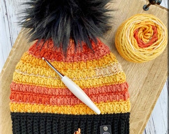 May include: A crocheted hat with a black pom pom and stripes of orange, yellow, and cream. The hat has a black band at the bottom and a small black label with a white bird logo. A crochet hook and a ball of yarn are also in the image.