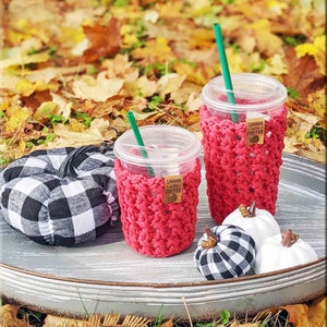 May include: Two red crocheted coffee cup cozies with the words "Shhh... Coffee" on a brown tag. The cozies are on a grey tray with two black and white checked pumpkins and two white pumpkins.