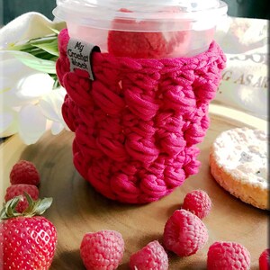 Peut inclure: Un manchon de tasse en crochet rose avec le texte "My Crochet Shack" sur le côté. Le manchon est sur une tasse en plastique transparente avec une boisson rose à l'intérieur. Il y a des framboises et une fraise sur une surface en bois.