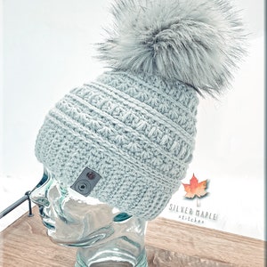 K&ouml;nnte beinhalten: Eine hellgraue H&auml;kelm&uuml;tze mit einem flauschigen wei&szlig;en Bommel. Die M&uuml;tze hat ein strukturiertes Muster und ein kleines braunes Lederlabel mit einem Ahornblatt-Logo, auf dem "Silver Maple Stitches" steht.
