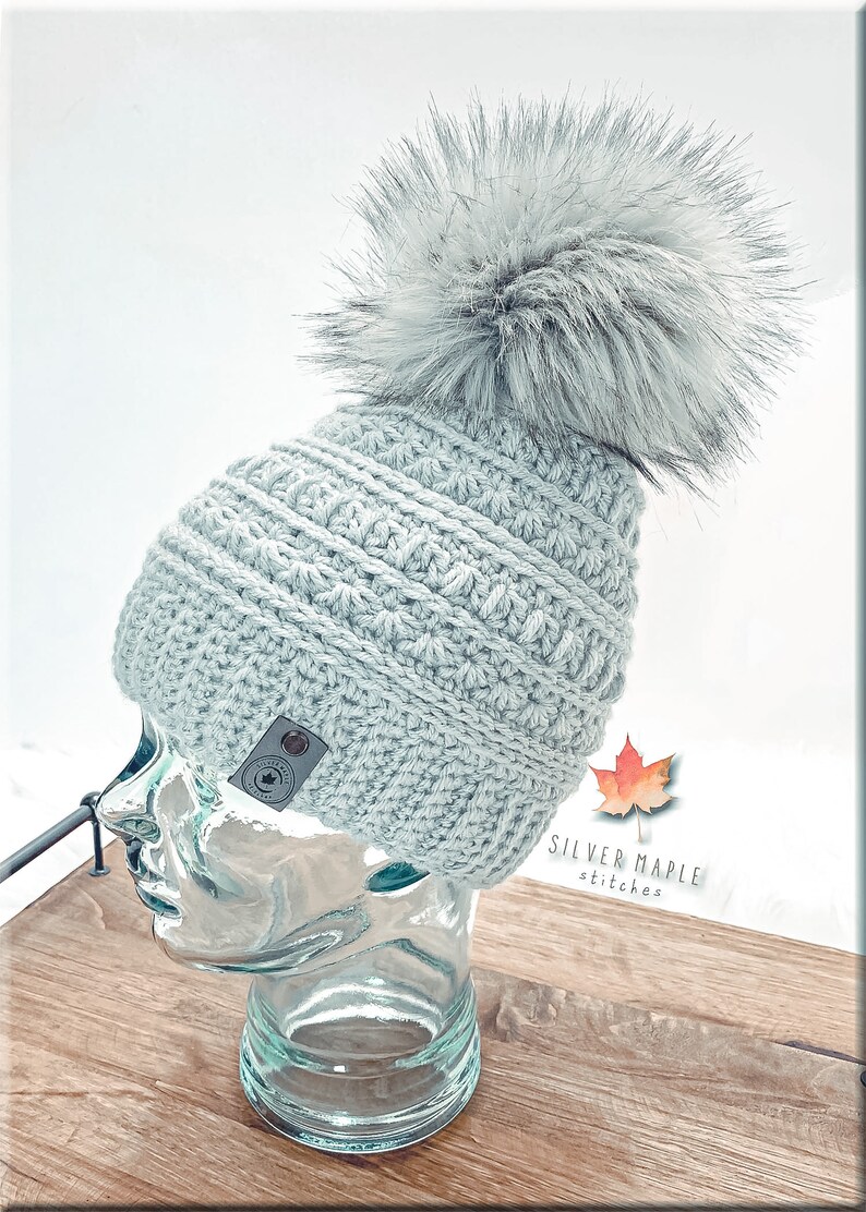 K&ouml;nnte beinhalten: Eine hellgraue H&auml;kelm&uuml;tze mit einem flauschigen wei&szlig;en Bommel. Die M&uuml;tze hat ein strukturiertes Muster und ein kleines braunes Lederlabel mit einem Ahornblatt-Logo, auf dem "Silver Maple Stitches" steht.