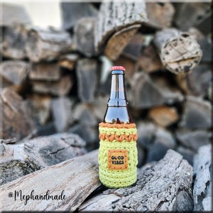 May include: A brown glass bottle with a green and orange crocheted koozie. The koozie has a brown leather patch with the text "GOOD VIBES" on it. The bottle is sitting on a pile of wood.