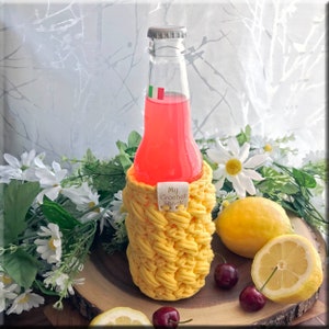 Op de afbeelding: Een gele gehaakte flessenwarmer met de tekst "My Crochet Shack" op een bruin houten oppervlak met citroenen en kersen.