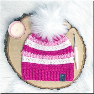 K&ouml;nnte beinhalten: Eine gestrickte M&uuml;tze in rosa und wei&szlig; mit einem wei&szlig;en Bommel oben. Die M&uuml;tze liegt auf einer Holzscheibe mit einer H&auml;kelnadel und einer Kerze.