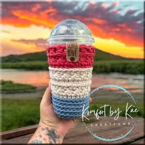 May include: A crocheted cup cozy in red, white, and blue colors. The cozy is made with a textured stitch and has a brown leather tag with the text "COFFEE LOVE".