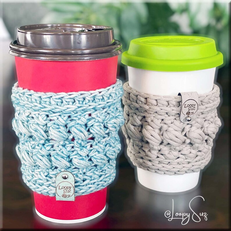 K&ouml;nnte beinhalten: Zwei Kaffeetassen mit geh&auml;kelten Bechern. Die Tasse links ist rot mit einem hellblauen Becher. Die Tasse rechts ist wei&szlig; mit einem beigefarbenen Becher. Beide Becher haben ein braunes Leder-Etikett mit dem Text "Loopy Suz" darauf.