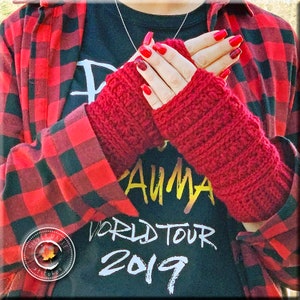 Puede incluir: Guantes sin dedos de crochet rojo con textura acanalada. Los guantes son usados por una persona que lleva una camisa de cuadros rojos y negros y una camiseta negra con el texto "Trauma World Tour 2019".