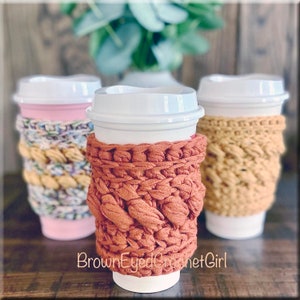 May include: Three crocheted coffee cup cozies in different colours and patterns. The cozies are made with a chunky yarn and have a textured look. The cozies are on top of white coffee cups. The text "BrownEyedCrochetGirl" is visible at the bottom of the image.
