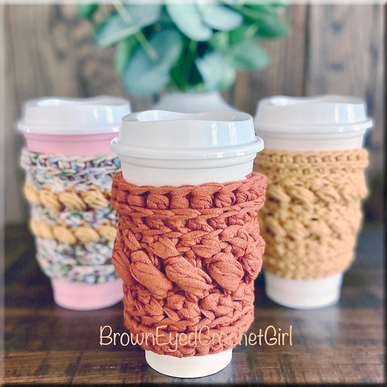 K&ouml;nnte beinhalten: Drei geh&auml;kelte Kaffeetassen-Cozies in verschiedenen Farben und Mustern. Die Cozies sind aus einem dicken Garn gefertigt und haben eine strukturierte Optik. Die Cozies befinden sich auf wei&szlig;en Kaffeetassen. Der Text "BrownEyedCrochetGirl" ist am unteren Rand des Bildes sichtbar.