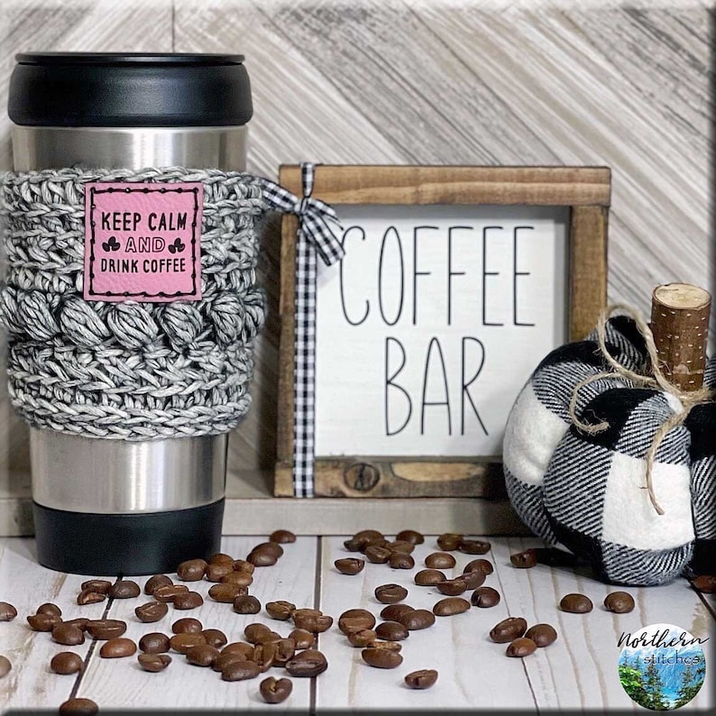K&ouml;nnte beinhalten: Ein grauer und wei&szlig;er geh&auml;kelter Kaffeetassenw&auml;rmer mit einem rosa quadratischen Patch, auf dem "Keep Calm and Drink Coffee" steht. Der W&auml;rmer befindet sich auf einer Edelstahl-Reise-Thermoskanne. Es gibt ein Holzschild mit der Aufschrift "Coffee Bar" und einen schwarz-wei&szlig; karierten K&uuml;rbis auf einer wei&szlig;en Holzoberfl&auml;che mit verstreuten Kaffeebohnen.