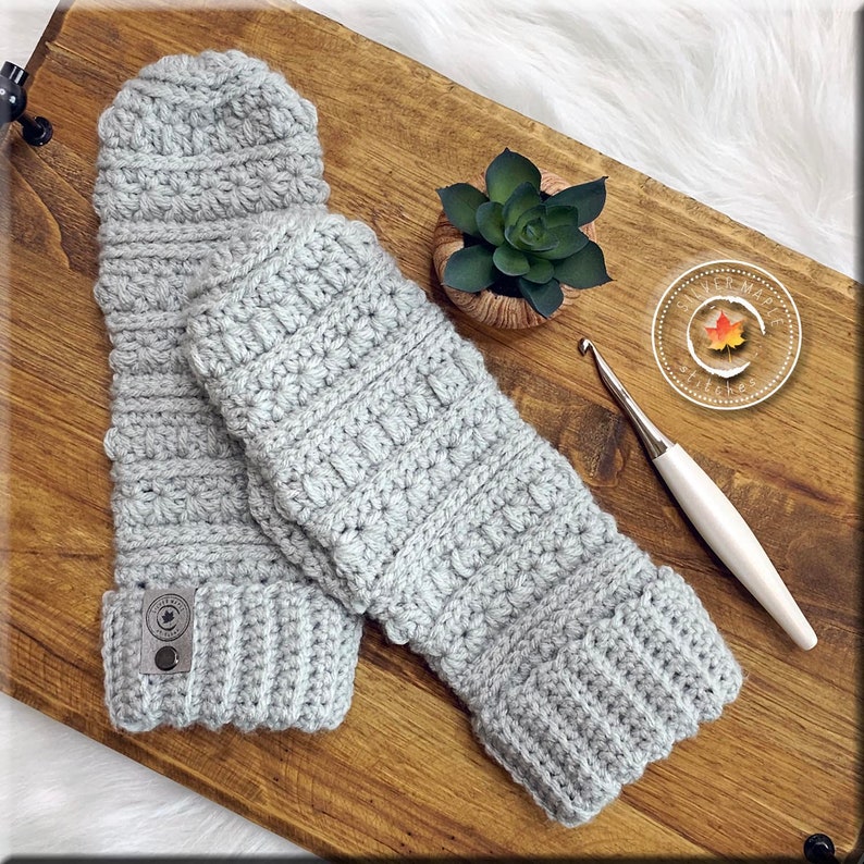 K&ouml;nnte beinhalten: Ein Paar graue, geh&auml;kelte Fingerlose Handschuhe mit einem strukturierten Stichmuster. Die Handschuhe liegen auf einer Holzoberfl&auml;che mit einer kleinen Sukkulente im Topf und einer H&auml;kelnadel.