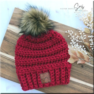 May include: Red crocheted beanie with a faux fur pom pom and a brown leather patch with the text "Smiz Silver Maple Stitches".