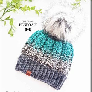 May include: A teal, gray, and white crocheted beanie with a fluffy white pom pom. The beanie has a brown leather tag with the initials "KJK" on it. The beanie is made by Kendra.K.