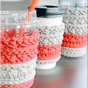 May include: Three crocheted cup cozies in pink and gray. The cozies are made with a chunky yarn and have a striped pattern. The cozies are on a table with a gray surface. The text "@LoopySuz" is visible in the bottom right corner of the image.