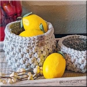May include: Two crocheted baskets in a light gray color, one with a tag that says "Love is love" and the other with yellow lemons inside. The baskets are on a wooden surface with white berries.