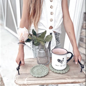 May include: A wooden tray with two green crocheted coasters and a white mug with a black rooster design and the text "wake up call".