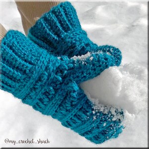 K&ouml;nnte beinhalten: Ein Paar tealblaue H&auml;kelhandschuhe mit einem strukturierten Muster. Die Handschuhe werden in einer Hand gehalten und ber&uuml;hren Schnee.