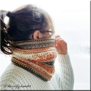 May include: A crocheted infinity scarf with stripes of brown, beige, peach, and white yarn. The scarf is worn by a person with long brown hair.