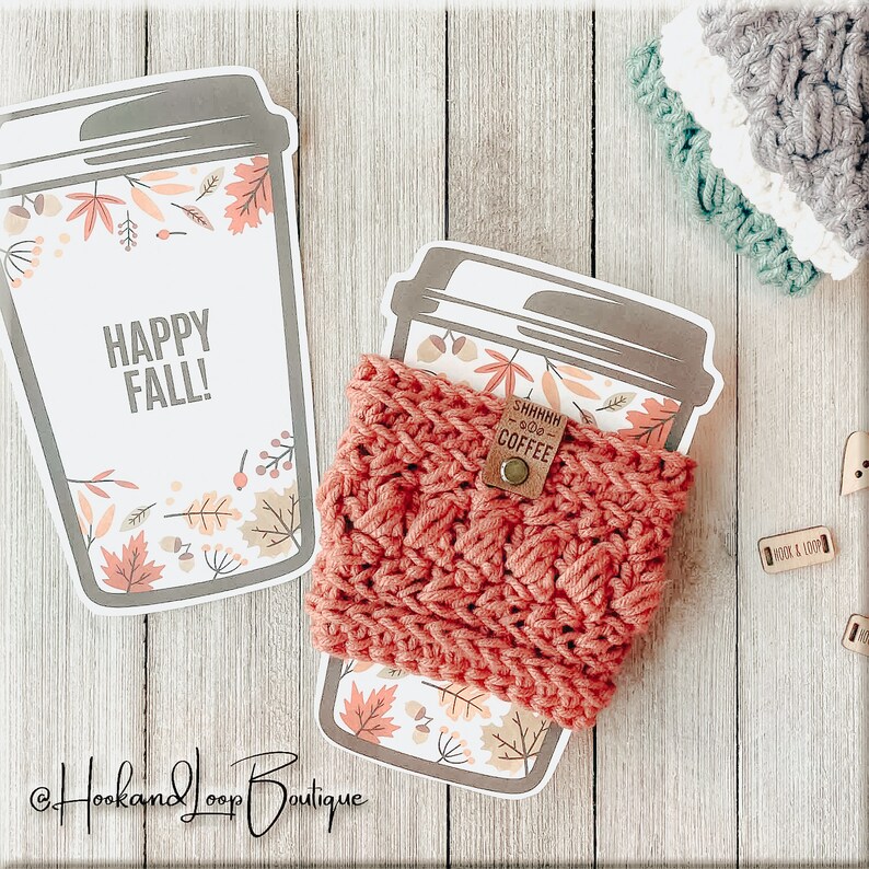 K&ouml;nnte beinhalten: Ein geh&auml;keltes Kaffeetassen-Cozy in Korallenfarbe mit einem braunen Lederanh&auml;nger, auf dem "Shhhh...Good Coffee" steht. Das Cozy liegt auf einem Papierkaffeebecher mit einem Herbstmotiv, auf dem "Happy Fall!" steht.