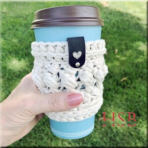 K&ouml;nnte beinhalten: Ein wei&szlig;er geh&auml;kelter Kaffeetassenw&auml;rmer mit einem schwarzen Lederriemen und einem silbernen Herzanh&auml;nger. Der W&auml;rmer befindet sich auf einer blauen und wei&szlig;en Pappkaffeetasse.
