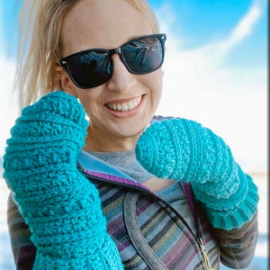K&ouml;nnte beinhalten: Eine Frau mit Sonnenbrille und einer gestreiften Jacke h&auml;lt ein Paar tealblaue, geh&auml;kelte Fingerhandschuhe. Die Handschuhe haben ein strukturiertes Stichmuster. Das Bild enth&auml;lt den Text "@lynzelow406".